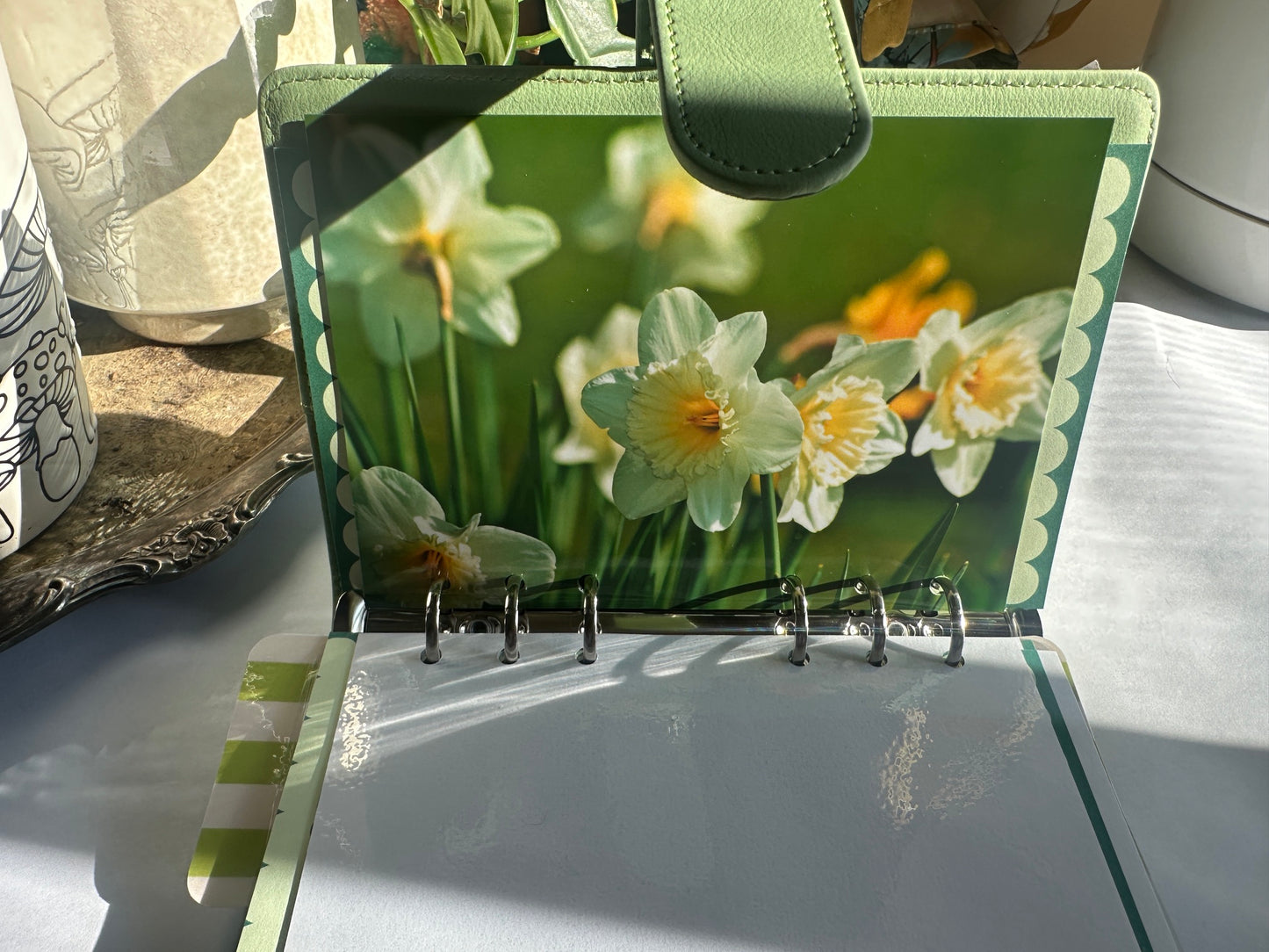 A green ring-bound planner or binder with a daffodil floral cover, shown open on a tabletop with sunlight and shadows.